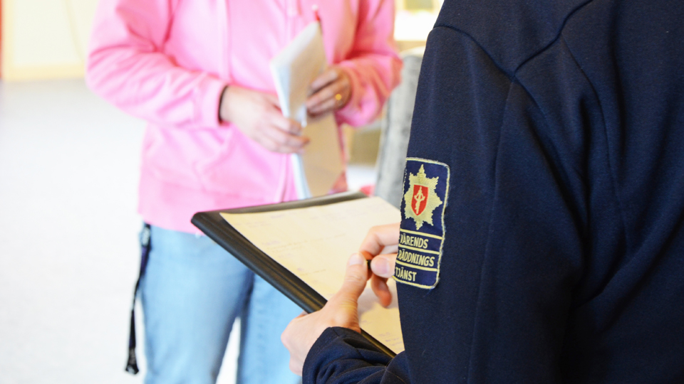 Person i räddningstjänstuniform håller i ett protokoll och en penna och samtal med en civil person, foto